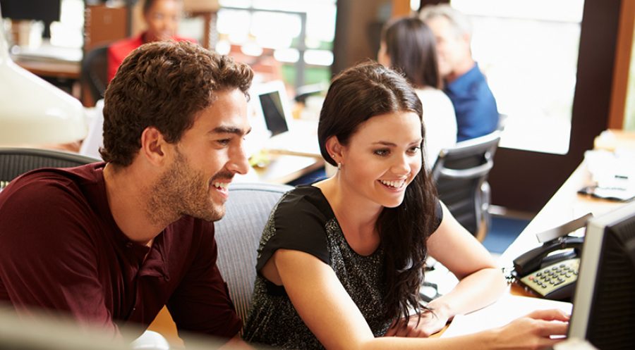 Two Colleagues Working At Desk With Meeting In Background